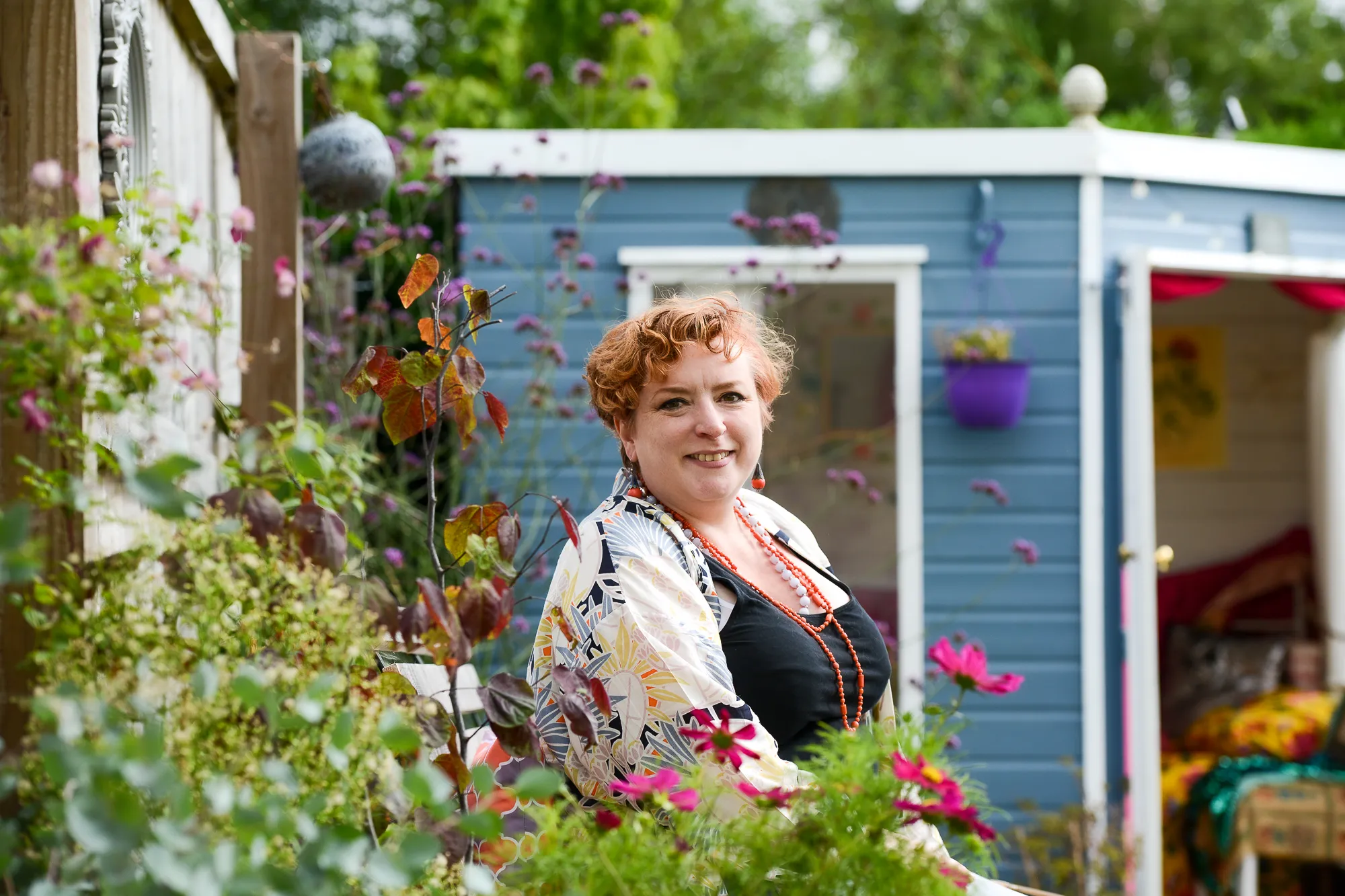 Charlotte Howard sitting on a bench in her garden with a summer house in the distance and a border in the foreground. Photo by Victoria Tapper