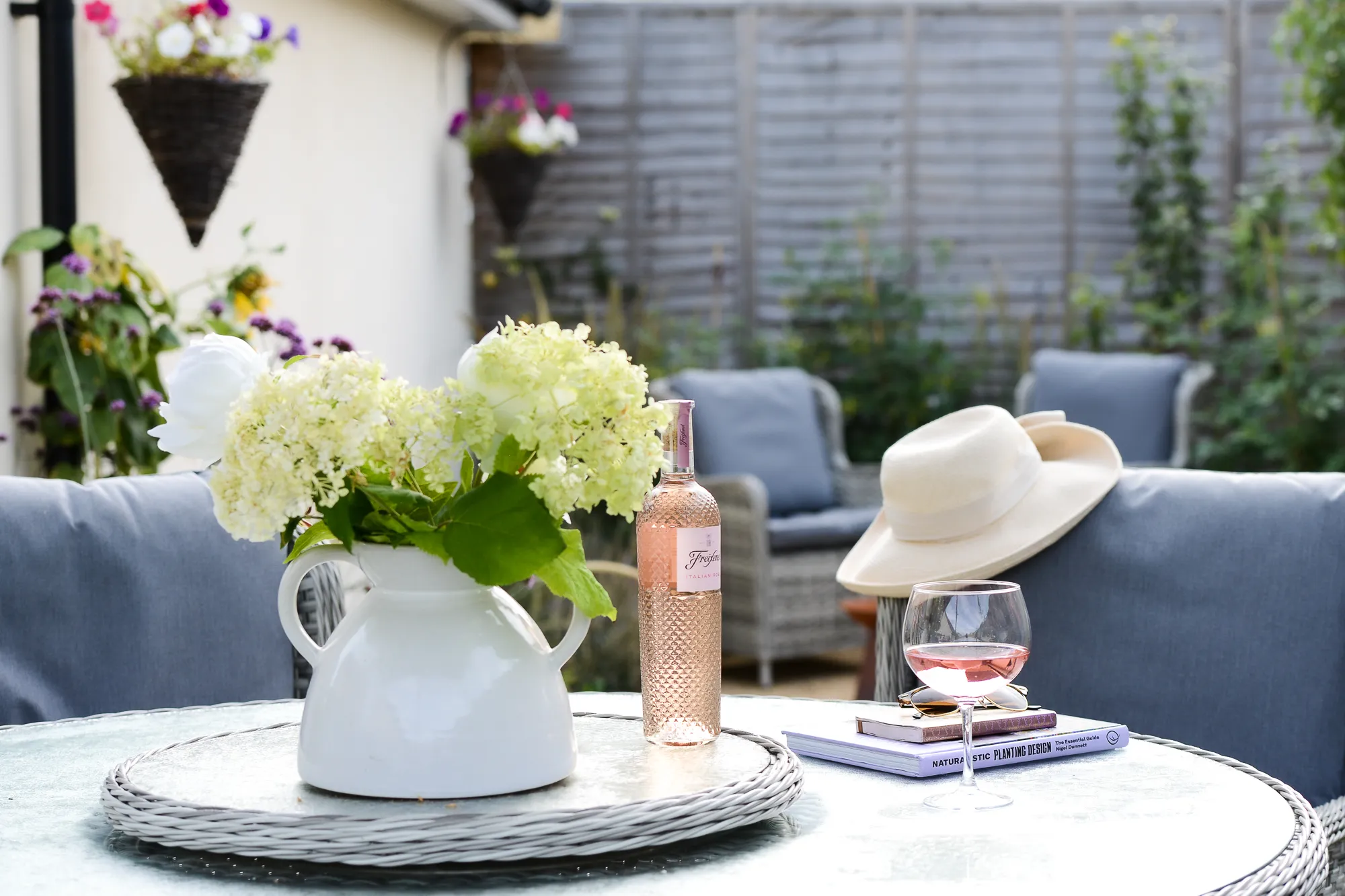 A bottle of wine, a glass and several books sit on a table in a garden. Photo by Victoria Tapper